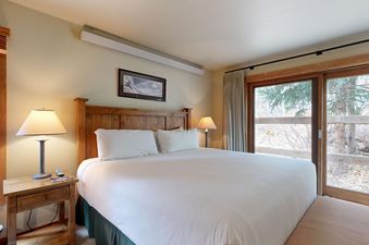 A cozy bedroom with a wooden king bed and sliding glass doors opening to outdoor pine trees.