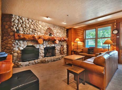 A cozy living room in Comstock 203 featuring a large stone fireplace, comfortable leather sofas, and views of green trees outside large windows.