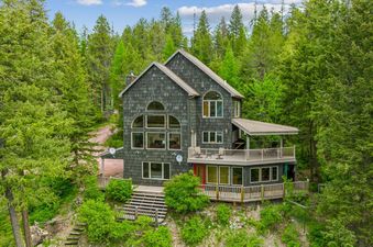 Elk Run, a dark grey shingled house, is nestled among lush green evergreen trees under a blue sky.