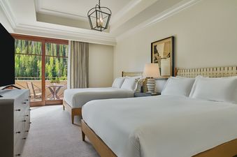 A spacious bedroom features two queen beds with white linens, a bedside table with a lamp, a dresser with a television, and a balcony overlooking a lush green forest.