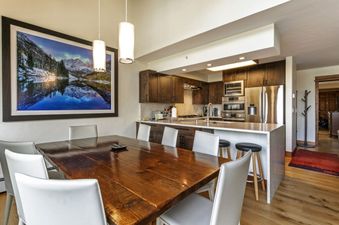 A dining room with a large table and chairs in front of a kitchen with stainless steel appliances and a large window overlooking a snowy mountain.