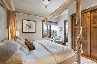 A cozy bedroom with a large bed, featuring light-colored linens, several decorative pillows, and an artistic headboard. A window with an outdoor view and a framed art piece are visible.