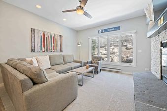 A cozy living room in The Charter A2505 with a large sectional sofa, a fireplace, and a view of the outdoors through big windows.