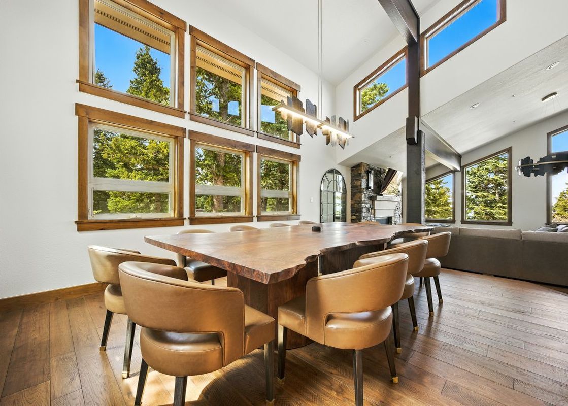 A spacious dining area featuring a large live-edge wooden table, leather chairs, and multiple windows offering views of evergreen trees.