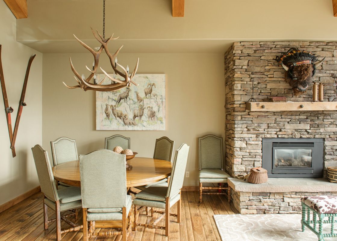 A rustic dining area with a round table and an antler chandelier, adjacent to a stone fireplace.