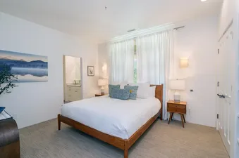 A bright bedroom with a queen-sized bed, two nightstands with lamps, a large window with white curtains, and a door leading to another room.