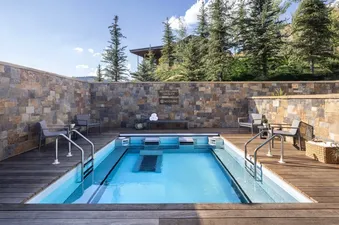 An outdoor pool surrounded by a wooden deck and stone walls, with pine trees and a distant building in the background under a clear sky.