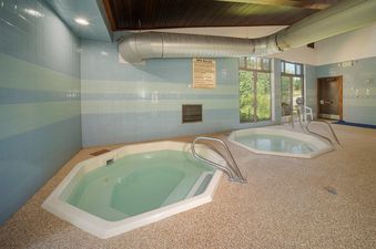 A spa area with two hot tubs, light blue and light green tiled walls, and a large window looking out onto trees.