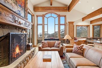 A cozy living room with a stone fireplace, leather sofas, and large windows offering a panoramic view of snow-covered mountains.