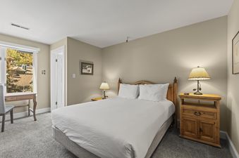 A cozy king bedroom at Fashion Coalition 402 features a wooden bed, two nightstands with lamps, a desk by the window, and soft grey carpeting.