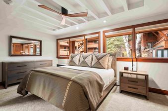 A king bedroom in Fasching Haus 9 and 10 featuring a large bed with decorative pillows and a throw blanket, a wooden dresser with a mirror, and expansive windows looking out onto a wooded area.
