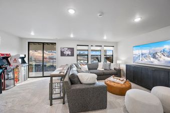 A modern and inviting living room featuring a large sectional sofa, a tufted ottoman, and a high-top table with stools, complemented by two arcade machines and large windows showcasing an exterior view.