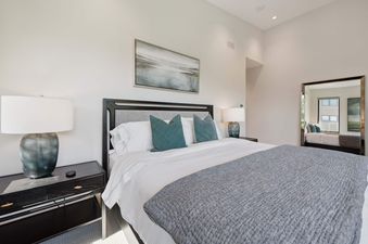 A serene bedroom in One Riverfront Townhome 07 featuring a bed with white linens, teal throw pillows, dark nightstands, and elegant lamps, with a landscape painting above the bed and a mirror reflecting a window.
