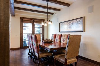 A rustic elegant dining room with a large wooden table, patterned chairs, a chandelier, and a framed winter landscape artwork on the wall.