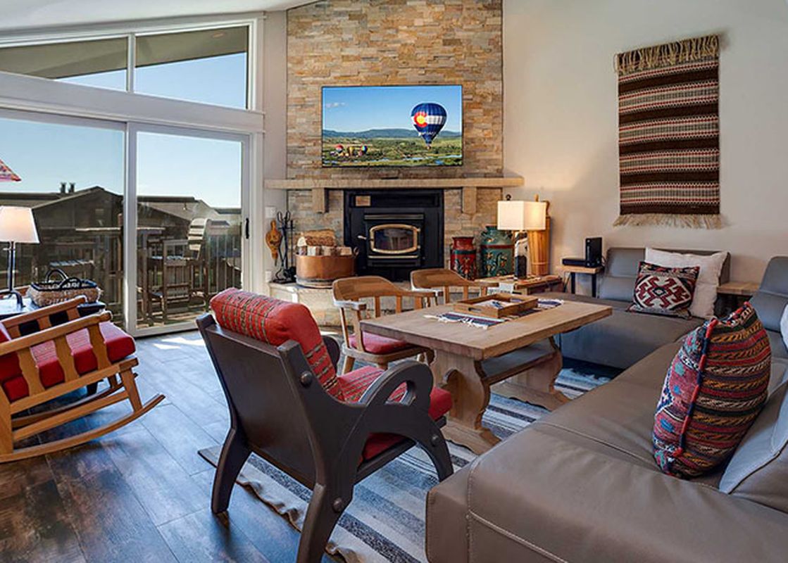 A cozy living room in Ranch at Steamboat RA220 features a gray sofa, a wooden dining table, a stone fireplace, and large windows with outdoor views.