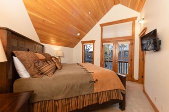 The primary bedroom at the Timber Trail Lodge offers a king-sized bed with rustic wood furnishings, a wall-mounted TV, and a private balcony.