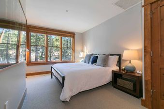 A bedroom with a large bed, a bedside table, a lamp, and a window looking out to the trees
