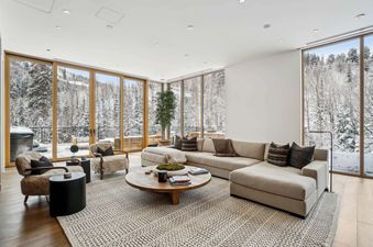 The Founders Place 1701 living room with an L-shaped sofa, armchairs, and a coffee table, all facing floor-to-ceiling windows with a snowy mountain view.