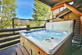 A bubbling hot tub on an outdoor deck at Comstock 203, surrounded by wooden railings, with a view of green trees and mountains under a blue sky.
