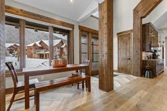 A rustic dining area with a wooden table, chairs, and a bench, facing large windows that offer a view of a snow-covered village.