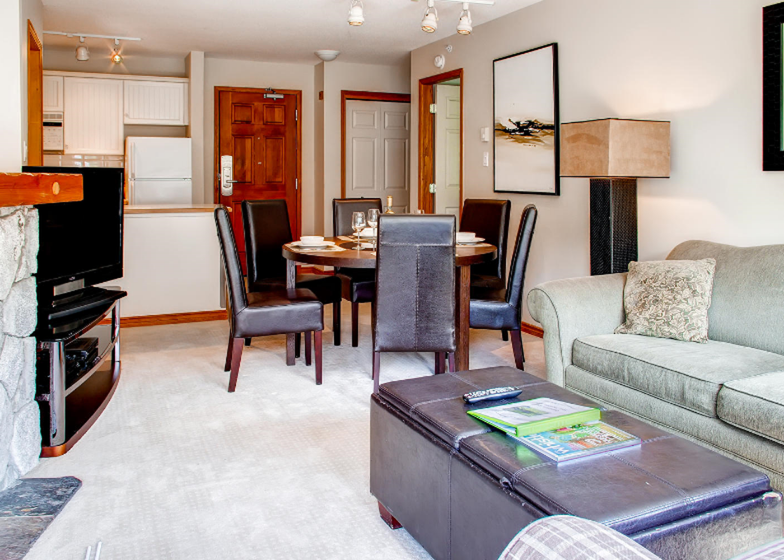 The dining area and living room of Aspens on Blackcomb Unit 547 are showcased, featuring a round wooden dining table with leather chairs and a comfortable sofa.