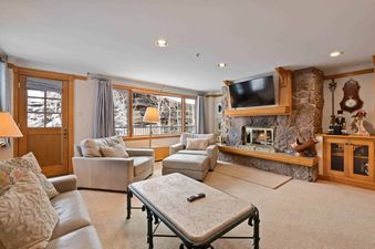 A cozy living room in Lodge Tower 387 3BR featuring a stone fireplace, a flat-screen TV, comfortable seating, and a large window with a view of a snowy winter landscape.