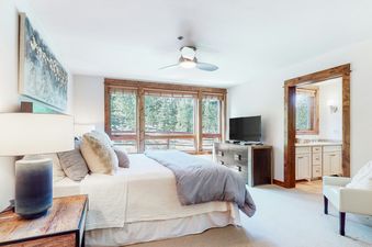 The BlueSky Breckenridge 504 bedroom features a large bed with neutral bedding, wood-framed windows offering a forest view, and a TV on a modern dresser.