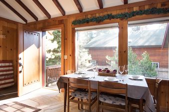 A rustic dining room with a set table, large windows overlooking trees and a cabin, and an open wooden door leading to a balcony.