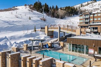 A luxurious ski-in/ski-out resort, Apex 1501, features an outdoor swimming pool and hot tubs set against a snow-covered mountain with active ski slopes and lifts under a clear blue sky.