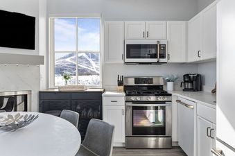 The image shows the modern kitchen and dining area of The Mason 2003 2005, featuring white cabinetry, stainless steel appliances, and a window overlooking snow-covered mountains.