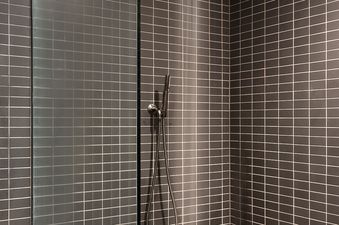A modern shower with dark grey tiles, a rainfall showerhead, and a small wooden stool.