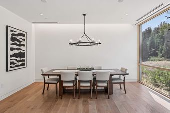 A modern dining room in Founders Place 1403 with a dark wood table, six upholstered chairs, a contemporary chandelier, and a large window offering a view of a forested landscape.