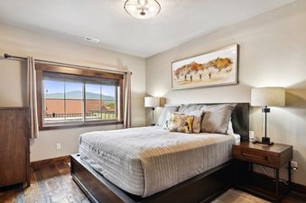 A master bedroom in Baker Loft 302 features a bed with soft bedding, decorative pillows, and wooden furniture, complemented by a large window overlooking mountains and rooftops.