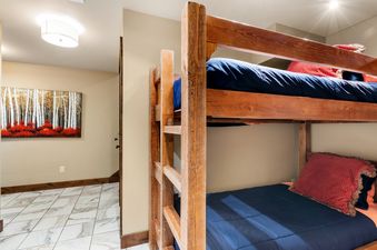 Two-tiered wooden bunk beds are made with navy blue comforters and pillows. In the background, there is a hallway with white and gray marble tile and a painting of birch trees with red leaves.