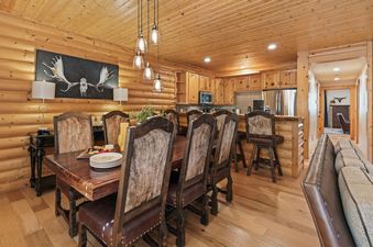 A rustic-chic dining area with a large wooden table and cowhide-upholstered chairs, illuminated by a modern chandelier, adjoins a fully equipped kitchen with light wood cabinets and stainless steel appliances.