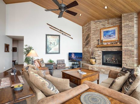 A cozy living room at Chateau Chamonix CX142 with a brick fireplace, comfortable seating, a wooden ceiling, and a television displaying a vibrant scene.