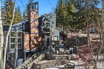 A modern chalet with dark siding and wooden accents stands among pine and aspen trees under a clear blue sky, featuring a terraced patio with a fire pit.