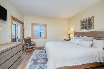 The inviting bedroom at Sundance Condos 202 features a large bed with a wooden headboard, soft bedding, a patterned area rug, and a comfortable armchair, all set in a warm, neutral-toned room.