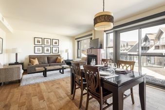 A spacious living room with a brown sofa and a dining area with a dark wood table set for dinner, all bathed in natural light from large sliding glass doors.