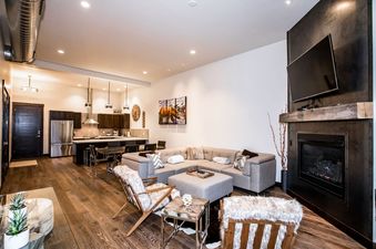 A spacious and modern open-concept living room and kitchen area with dark wood floors, a large gray sectional sofa, faux fur chairs, and a sleek fireplace with a mounted TV.