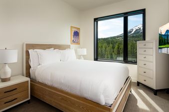 Moonlight Basin 212 Lakelodge is a bright, spacious bedroom featuring a king-size bed with white linens, a wooden dresser, and a large window with stunning mountain views.