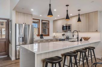A modern kitchen with light wood cabinets, stainless steel appliances, a white island, and three black pendant lights.