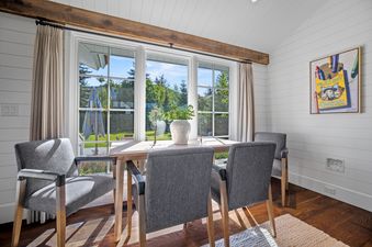 A bright dining area in Bluebell Cottage with a wooden table, four grey chairs, large windows with an outdoor view, white shiplap walls, and a rustic ceiling beam.