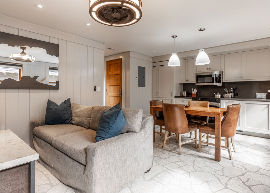 A comfortable open-concept living room and kitchen at Cedar Crest Chickadee Cottage 2, featuring a gray sofa, a wooden dining table with leather chairs, and modern kitchen appliances.
