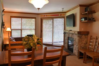 A cozy living space features a dining area with a wooden table, a stone fireplace, and large windows with blinds.