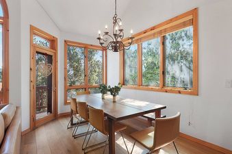 A wooden dining table with six chairs sits in the center of a room with large windows. The room has wooden floors, a white ceiling, and a chandelier hanging above the table.