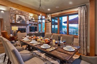 Dining room table with chairs, lit candles, and a bottle of wine set for a dinner party. The room features a large window with a mountain view in the background, a stone fireplace, and a rustic wooden table.