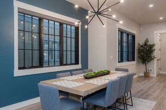 A modern dining room in Gather and Glow features a light wood table with eight grey upholstered chairs, black-framed windows, and a black sputnik chandelier.