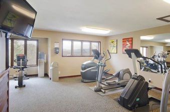 A well-lit fitness center with exercise equipment, large windows showing an outdoor view, and mirrors on one wall.