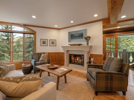 A cozy living room in Chateau Beaver Creek Residence 203 featuring a fireplace, comfortable seating, and a large arched window with a view of evergreen trees.